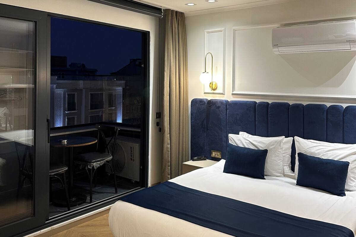 A modern hotel room with a large bed, a balcony with a table and chairs, and an air conditioning unit on the wall.