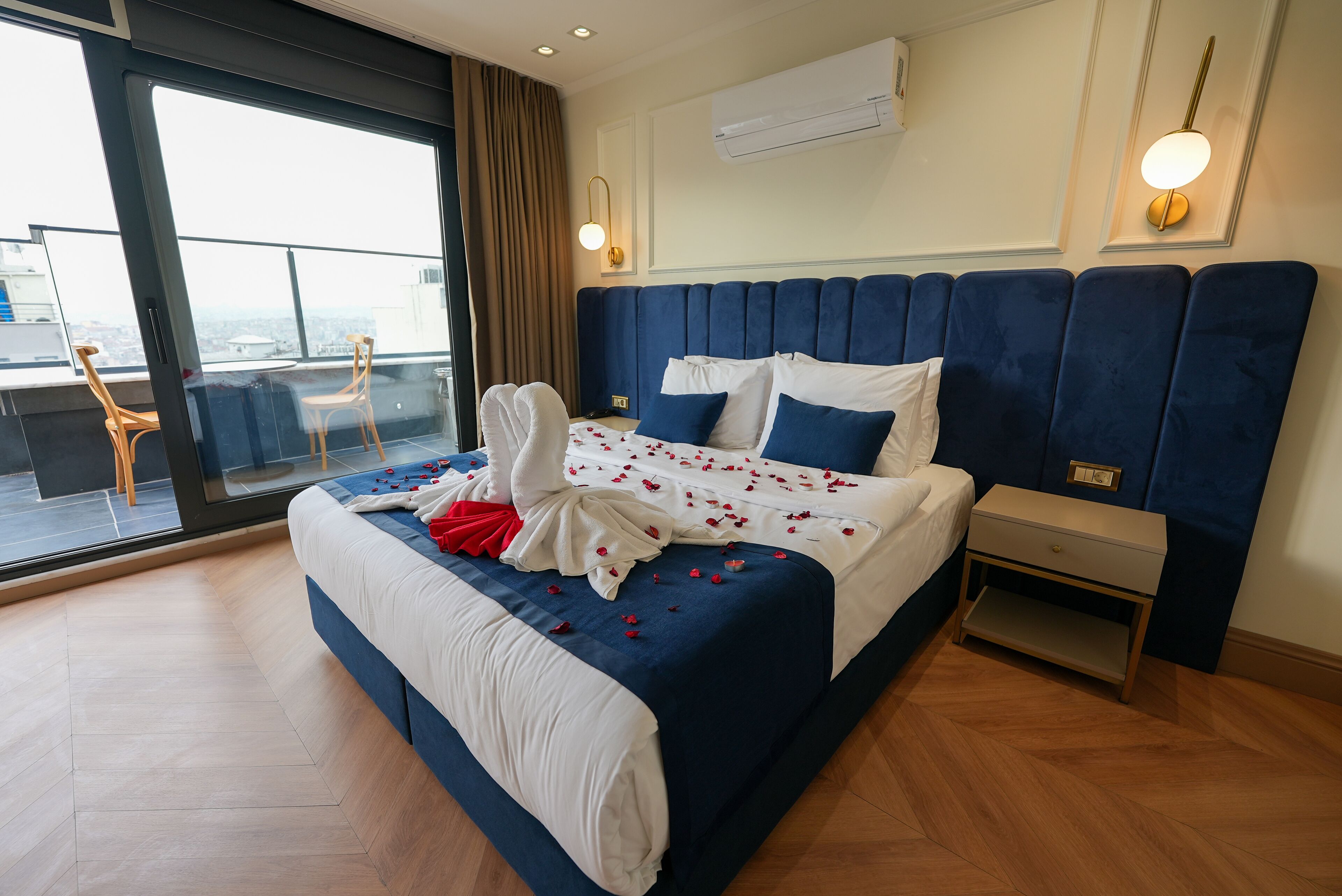 A hotel room with a large bed, a nightstand, a wall-mounted air conditioner, and a view of the city through a large window.
