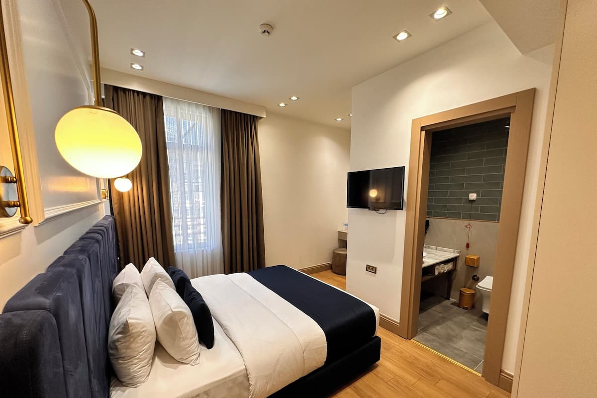 A modern hotel room with a bed, a TV mounted on the wall, a bathroom visible through an open door, and a window with curtains.