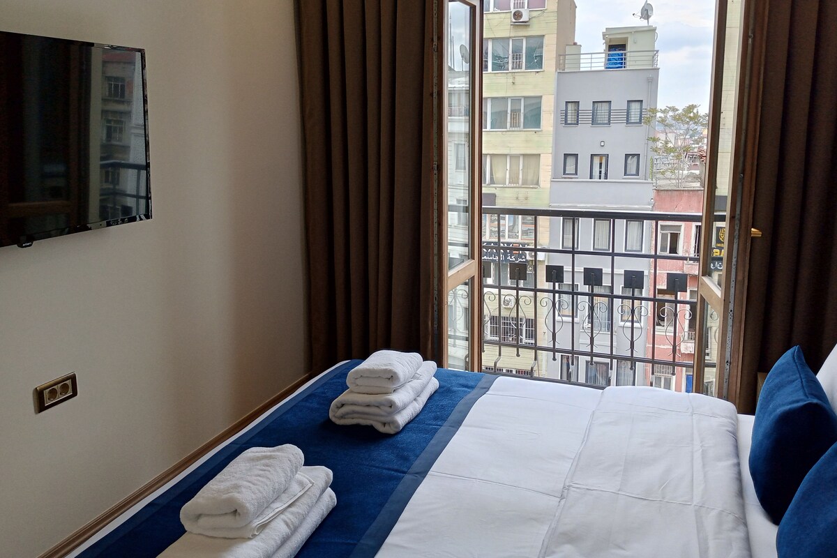 A hotel room with a bed, a TV mounted on the wall, and a view of the city through the window.