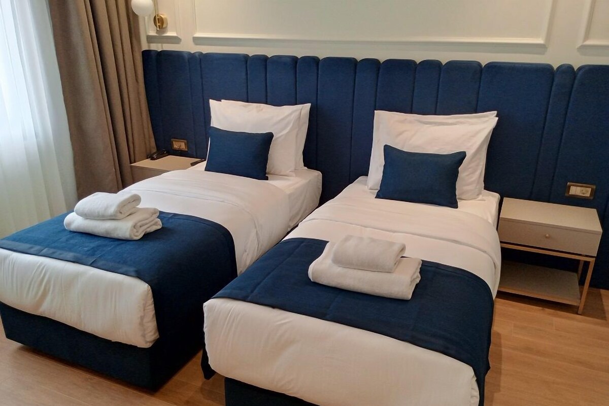 A hotel room with two beds, each with a blue headboard, white bedding, and a folded towel on the foot of each bed.