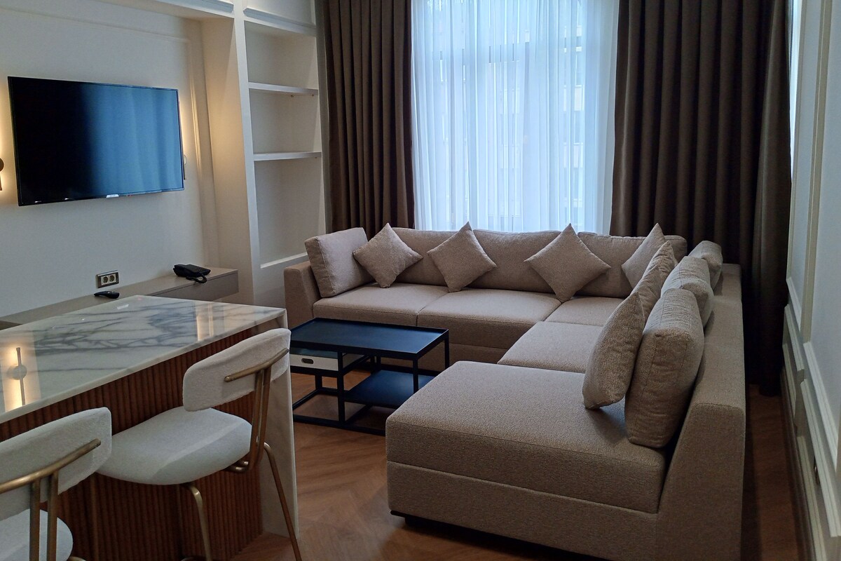 A modern living room with a sectional sofa, a flat-screen TV mounted on the wall, a dining area with a marble table and chairs, and a large window with curtains.