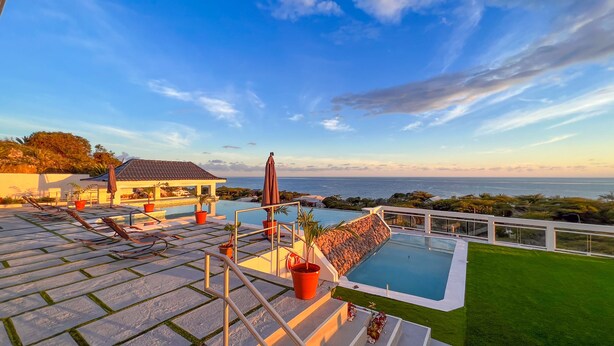 Un área de piscina lujosa con vista despejada al océano, un pabellón y un césped cuidado.