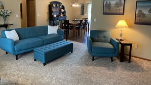 Ein Wohnzimmer mit einem blauen, modularen Sofa, dazu passendem Hocker, einem Sessel, einem Essbereich mit Tisch und Stühlen, einem Bücherregal und einer Lampe auf einem Beistelltisch.