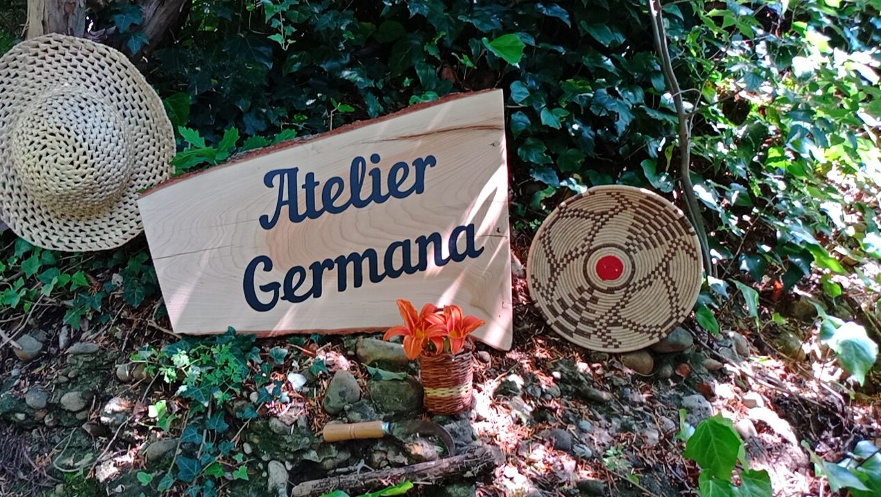 A wooden sign with the text "Atelier Germana" surrounded by natural elements like a woven basket, a woven hat, and a potted plant.