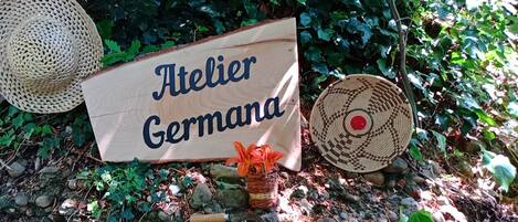A wooden sign with the text "Atelier Germana" surrounded by natural elements like a woven basket, a woven hat, and a potted plant.