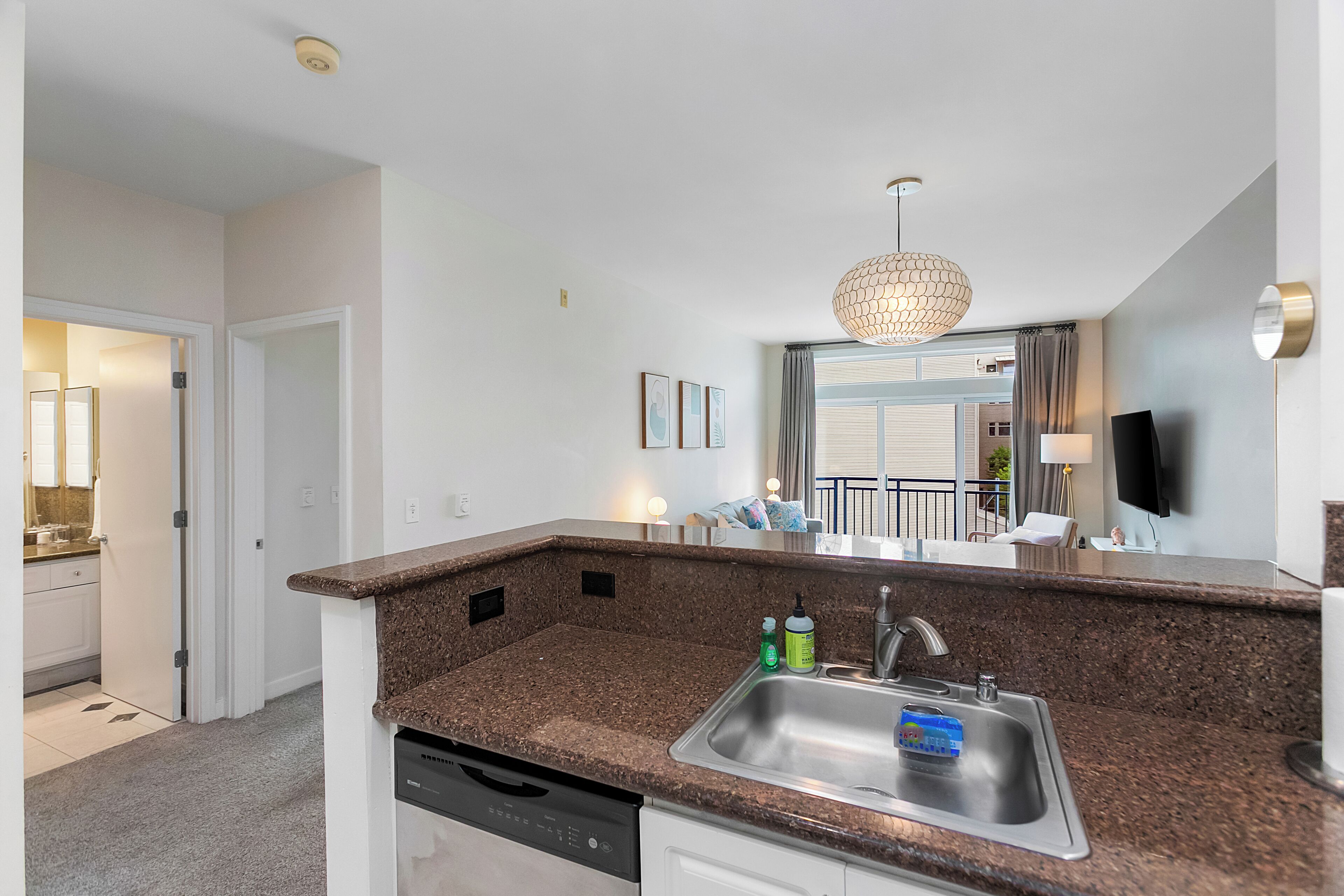 A modern kitchen with granite countertops, a stainless steel sink, and a dishwasher. There is a dining area with a table and chairs visible through an open door.