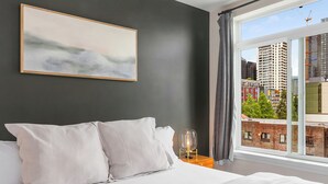 A bed with white linens, a nightstand with a clear glass lamp, and a cityscape view through a large window. - Belltown Perch explore like a local (Seattle)