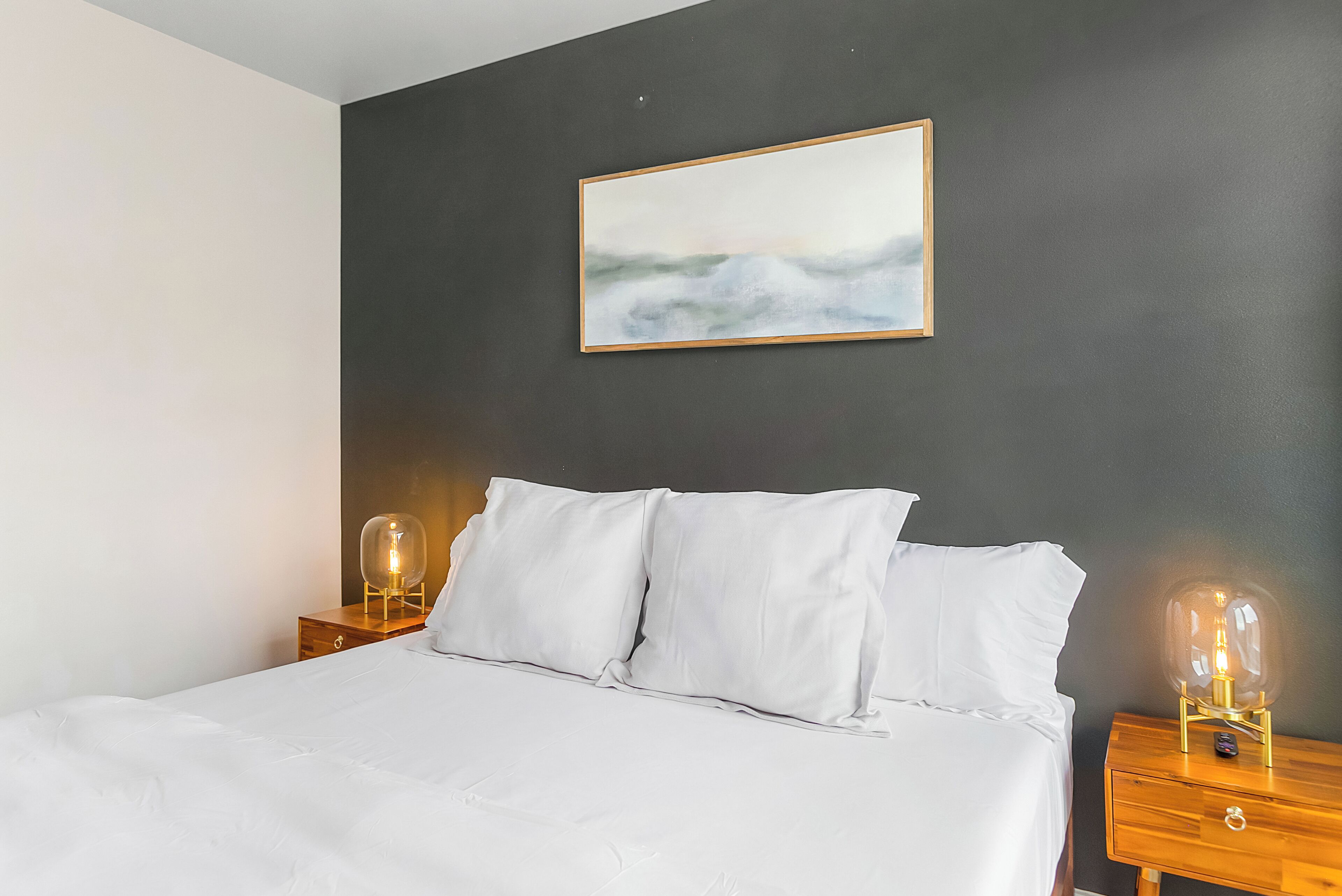 Een net opgemaakt bed met witte lakens, twee nachtkastjes met glazen lampen, en een ingelijst schilderij aan de donkere muur erboven.