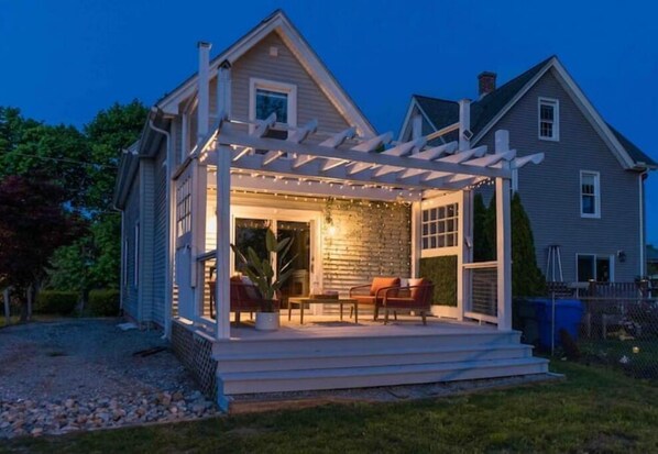 Relax on the covered porch with a seating area under a pergola, surrounded by a well-manicured lawn and a gravel driveway.