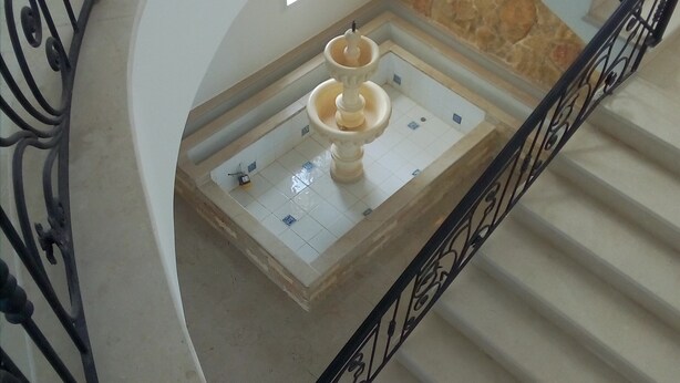 Eine geschwungene Treppe mit dekorativer Metallreling, einer Steinwand und einem kleinen Springbrunnen in der Mitte.