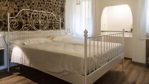 A bedroom with a white metal bed frame, a patterned tapestry on the wall, a bedside lamp, and a window with curtains. - Aponalview apartment  027042-LOC-12481 (Venezia)