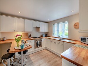 Une cuisine moderne avec des meubles blancs, un plan de travail en bois, une cuisinière à gaz, un évier et une fenêtre avec des stores.