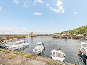 Un port esportiu amb diverses embarcacions amarrades, un moll de pedra i un turó amb edificis.