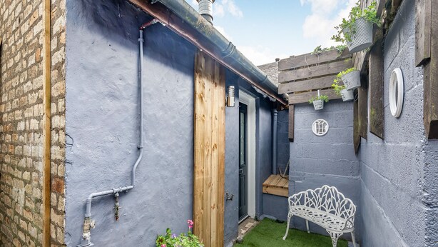 Un patio estrecho con una pared azul, una puerta de madera, un banco de metal blanco y una planta en maceta.