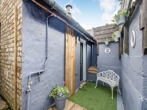 A narrow courtyard with a blue wall, a wooden door, a white metal bench, and a potted plant.