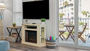 A living room with a fireplace, a flat-screen TV, a balcony with outdoor furniture, and a view of a building with palm trees. - Bikini Bottom *Destin Harbor* Private Gated Beach! (Destin)