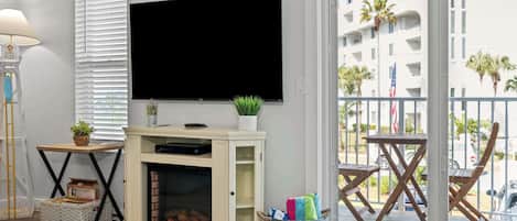 A living room with a fireplace, a flat-screen TV, a balcony with outdoor furniture, and a view of a building with palm trees.