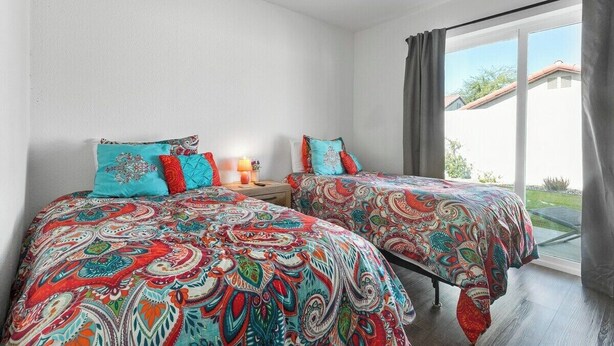 Une chambre avec deux lits, chacun agrémenté d’une literie aux motifs colorés, un ventilateur de plafond et une fenêtre avec des rideaux.