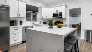 Une cuisine moderne avec des meubles blancs, un grand îlot central, des appareils électroménagers en acier inoxydable et un coin repas visible à travers une porte ouverte.