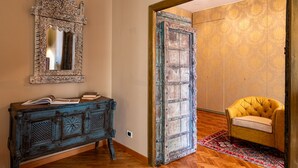 Ein Zimmer mit Holzboden, einem blauen, antiken Schminktisch, einem Spiegel mit aufwendigem Rahmen, einem gelben Sessel und einer teilweise geöffneten Tür, die in einen anderen Raum führt.