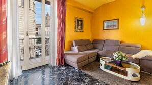 Ein Wohnzimmer mit einem Ecksofa, einem Glastisch mit einer Vase und Blick auf ein Gebäude durch ein mit Vorhängen drapiertes Fenster.