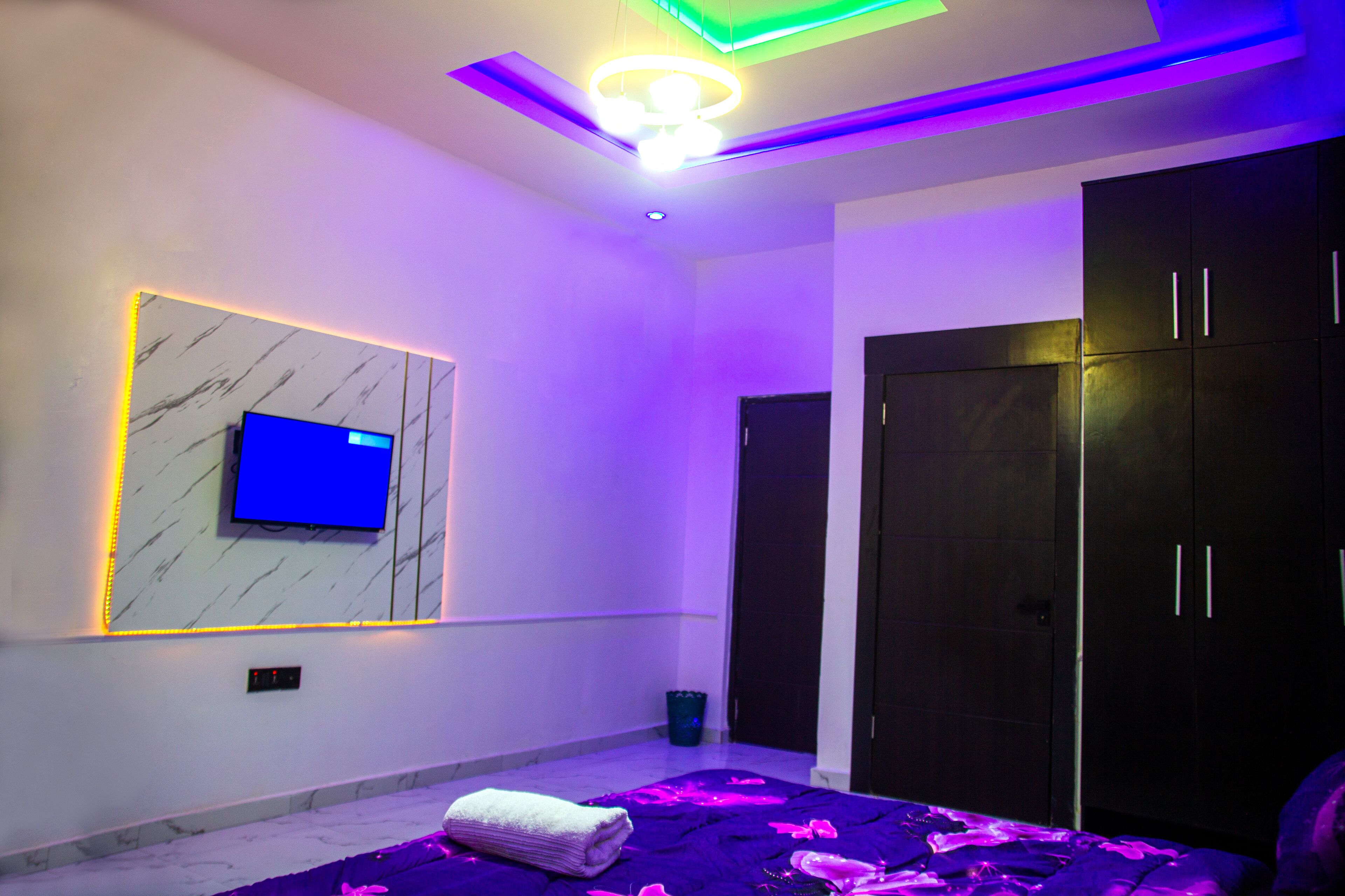 A modern bedroom with a TV mounted on a marble wall, a bed with a purple blanket, and a dark wardrobe.