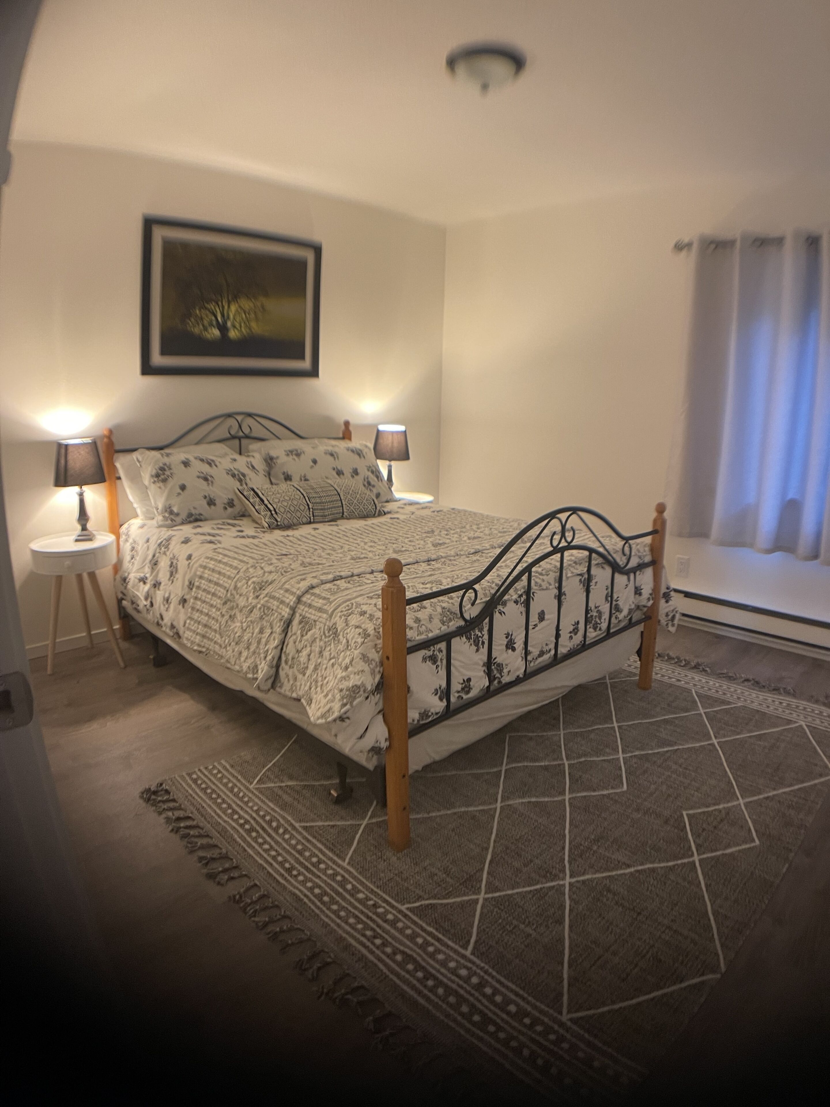 Ein Schlafzimmer mit einem Bett, zwei Nachttischen mit Lampen, einem Teppich, einem Bild an der Wand und einem Fenster mit Vorhängen.