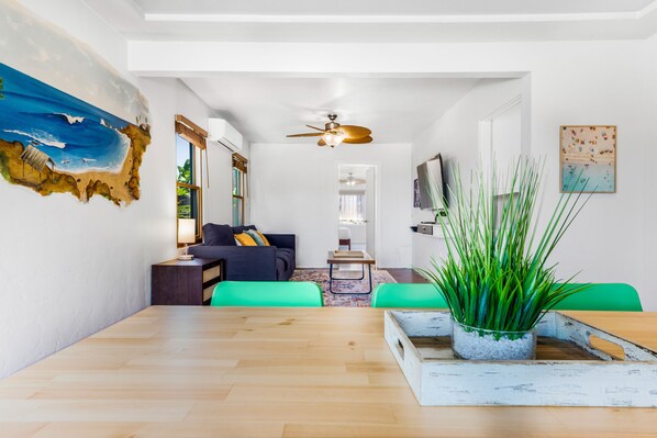 A modern living room with a wooden dining table, green chairs, a sofa with cushions, a ceiling fan, and a large painting of a beach scene.