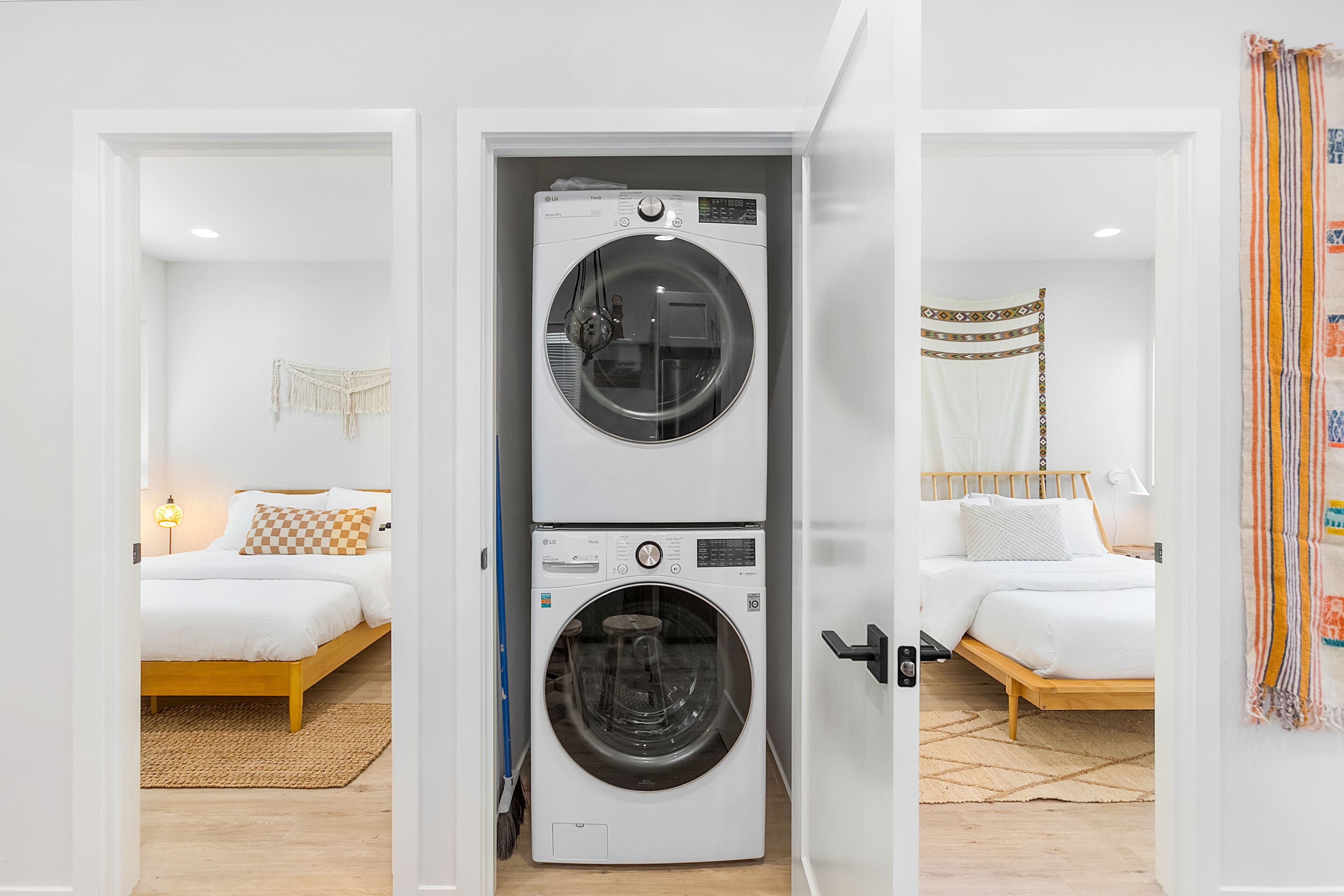 A laundry room with two stacked washing machines, a bed with white bedding, and a patterned throw on the bed.