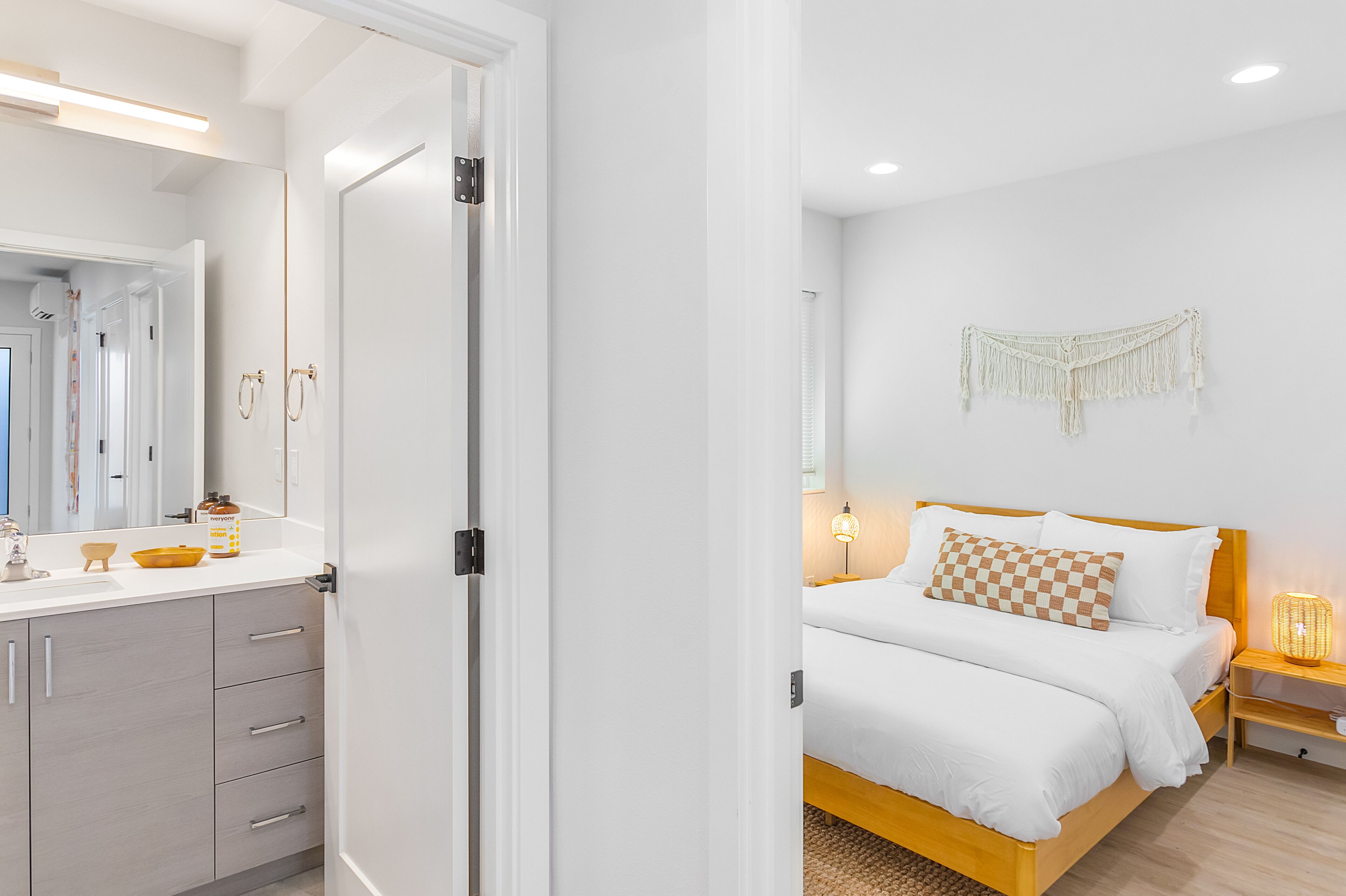 Un dormitorio moderno con una cama de madera, ropa de cama blanca, una mesita de noche con lámpara y un colgador de macramé en la pared.