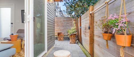 A narrow outdoor patio with wooden flooring, a sliding glass door, and a wooden fence. There are two wicker chairs and a small table with a plant.