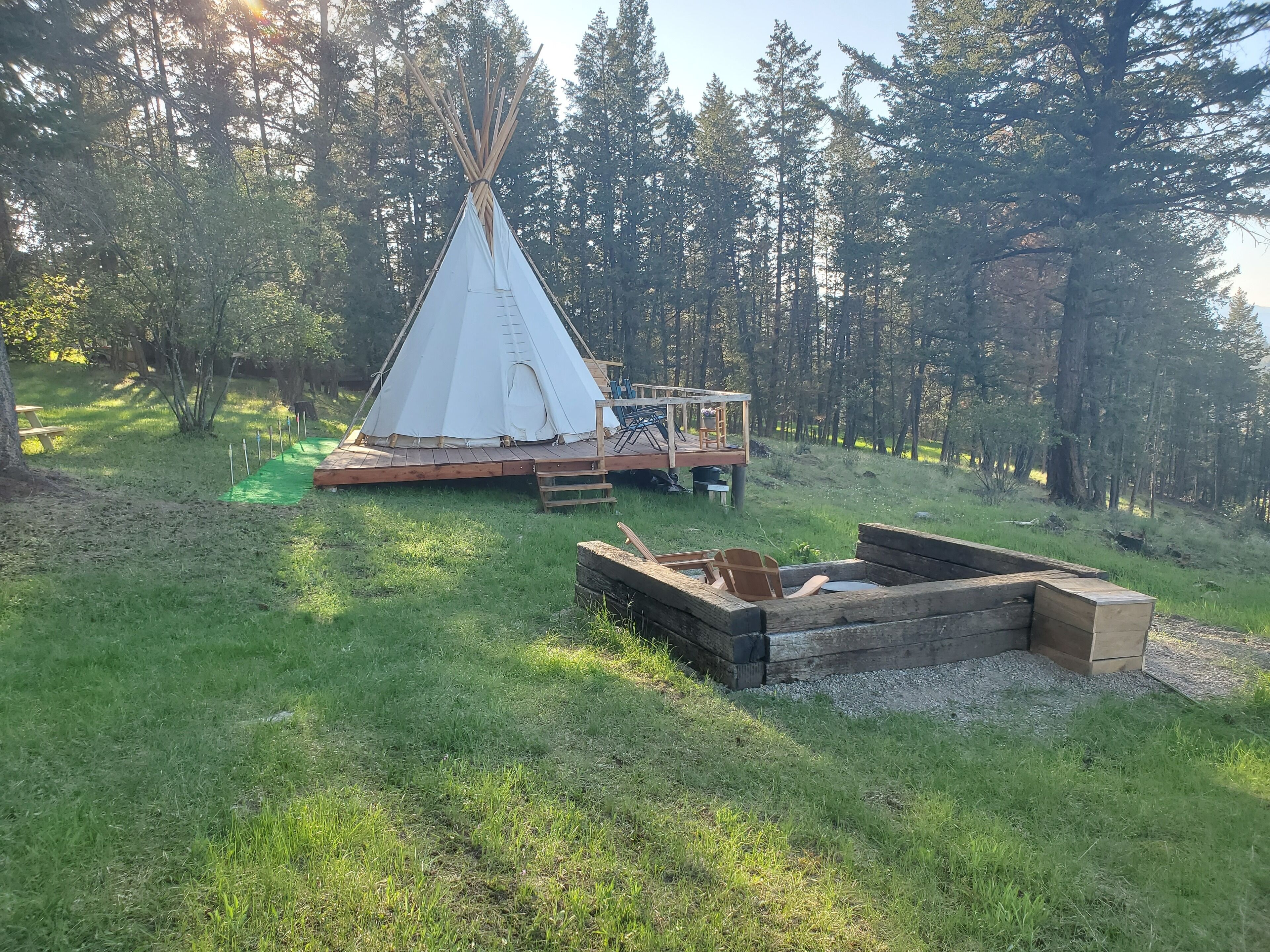 A teepee with a wooden deck and a fire pit in a forested area.