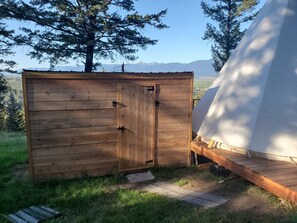 A wooden cabin with a door, a small porch, and a tent in a natural setting with mountains in the background. - Mountain Quest Tipi (Rexford)