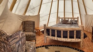 A cozy interior of a tent with a wooden bed, a patterned armchair, a small table, and a rug. - Mountain Quest Tipi (Rexford)