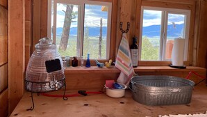 A rustic cabin interior with wooden walls, large windows, a metal bucket, a striped towel, and various cleaning supplies. - Mountain Quest Tipi (Rexford)
