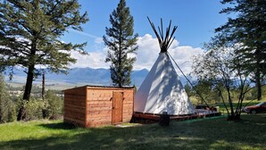 A wooden cabin and a white teepee in a grassy field with trees and mountains in the background. - Mountain Quest Tipi (Rexford)