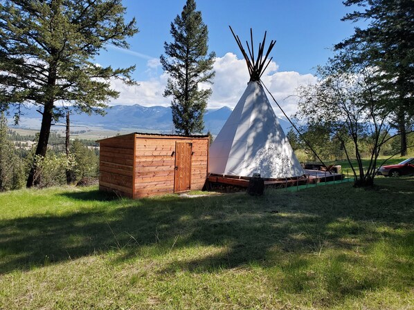 A wooden cabin and a white teepee in a grassy field with trees and mountains in the background. - Mountain Quest Tipi (Rexford)