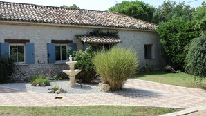 A stone building with a tiled roof, a small garden with a fountain, and a paved walkway.
