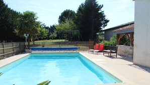 A rectangular swimming pool with a wooden deck surrounding it, a swing set, and a covered seating area with chairs.