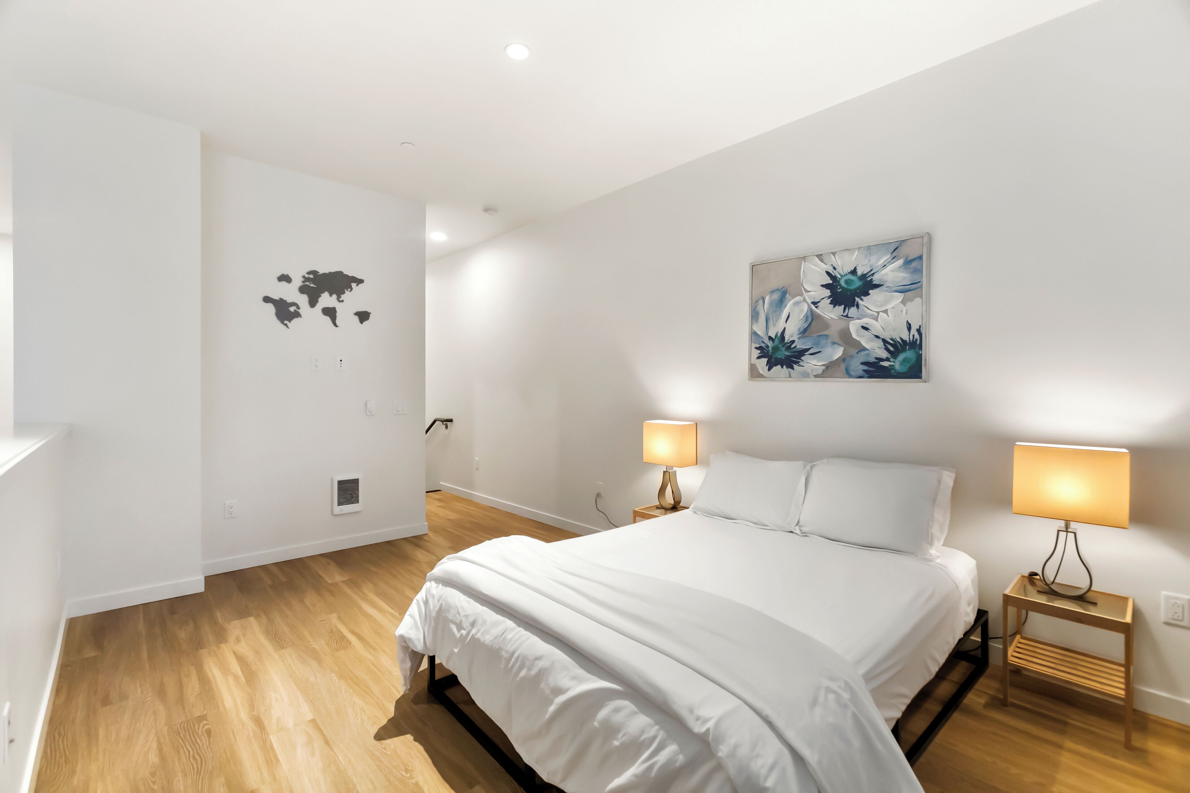 Un dormitorio con una cama grande, dos mesitas de noche con lámparas, un mapa mundial en la pared y un cuadro floral sobre la cama.