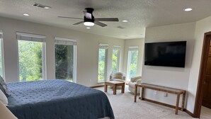 Een slaapkamer met een groot bed, een plafondventilator, een aan de muur bevestigde televisie en een raam met uitzicht op bomen.