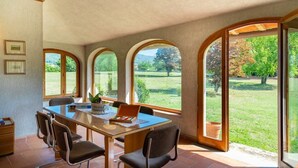 Uma área de refeições com mesa, cadeiras e grandes janelas em arco que oferecem vista para uma paisagem gramada.
