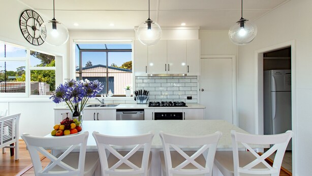 A modern kitchen with white cabinetry, a large island with bar stools, and pendant lighting. - Pheeney Place Five-star pet Friendly (Mallacoota)