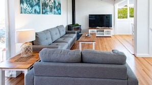 A modern living room with a grey sofa, a wooden coffee table, and a TV. There's a fireplace and a window with a view. - Pheeney Place Five-star pet Friendly (Mallacoota)