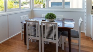 A dining area with a wooden table and white chairs, a potted plant, and a view of the outdoors through large windows. - Pheeney Place Five-star pet Friendly (Mallacoota)
