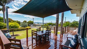 A wooden deck with a table and chairs, a covered area with a dark canopy, and a view of a green landscape with trees and hills in the background. - Pheeney Place Five-star pet Friendly (Mallacoota)