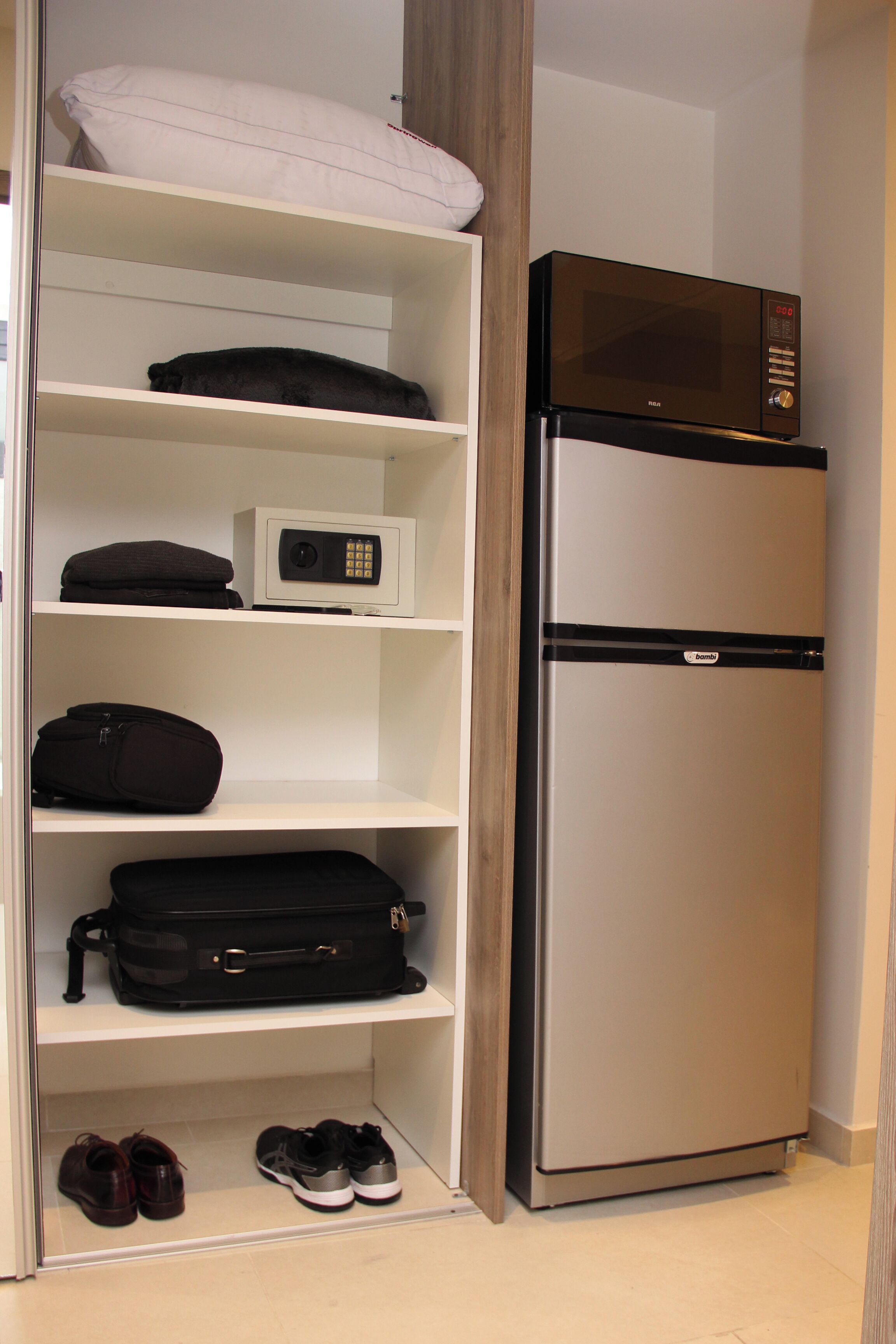 A compact room with a built-in microwave and refrigerator, a shoe rack, and a shelf with a pillow.