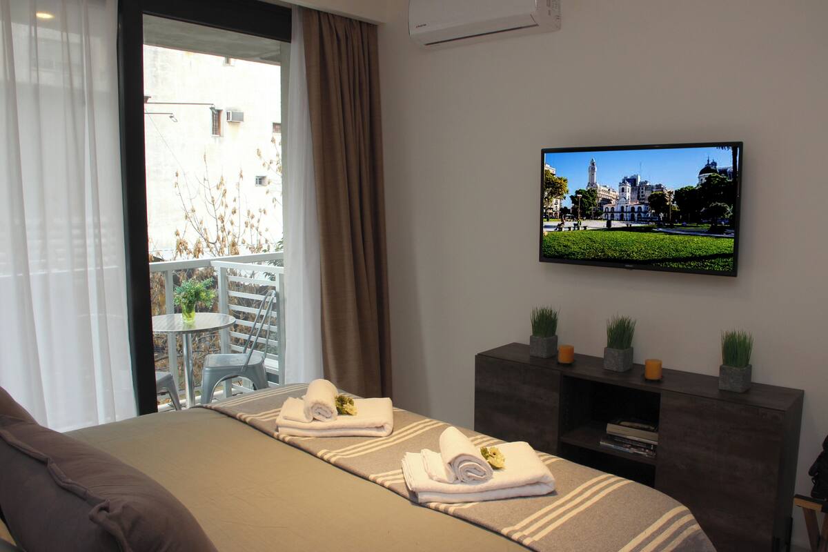 A hotel room with a bed, a TV mounted on the wall, a balcony with a table and chairs, and a view of a building outside.