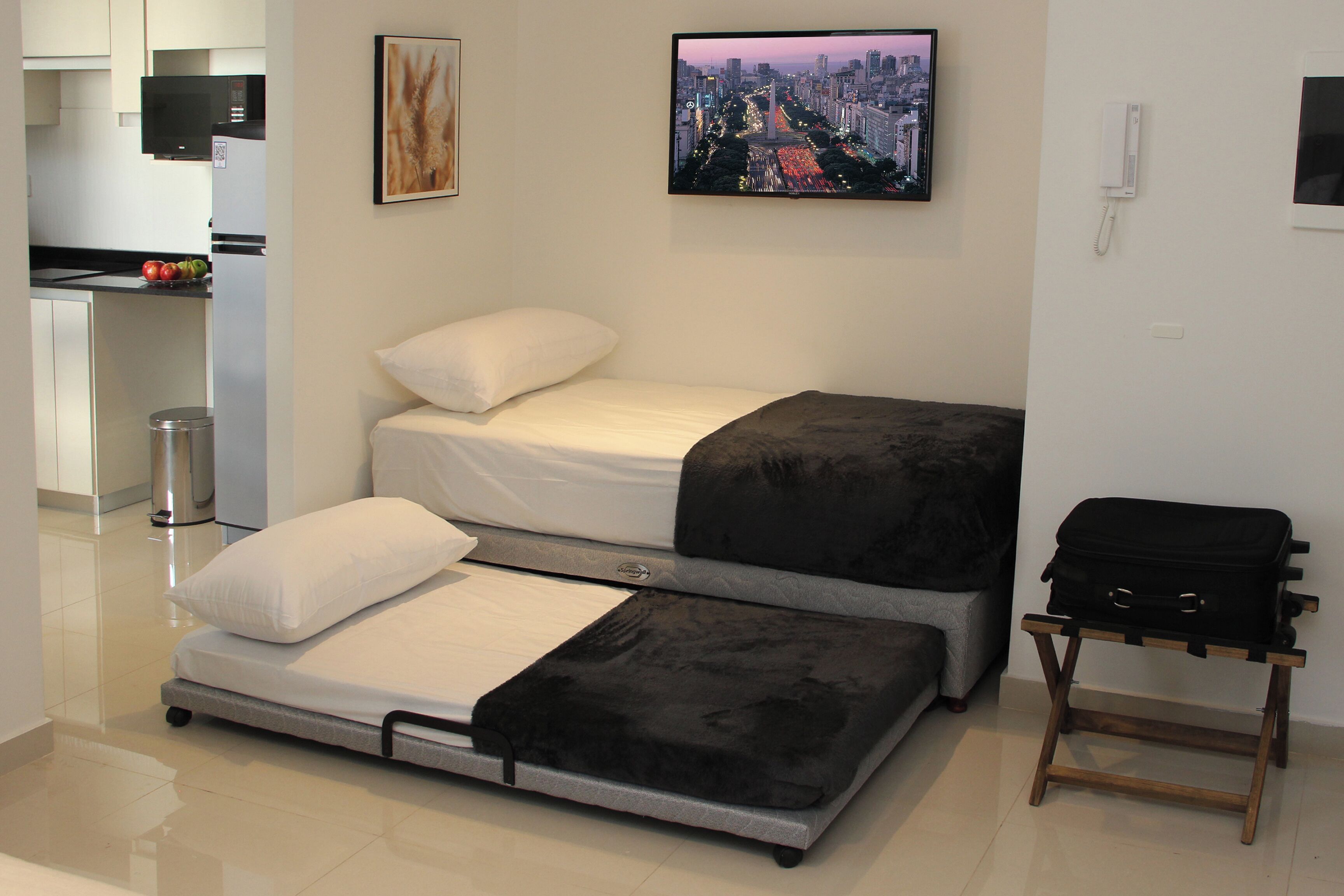 A compact room with a bed, a small table with a bag, a TV, and a kitchen area.